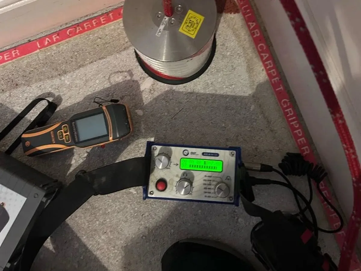 Ground microphone and blue acoustic amplifier with a lit display on a bare concrete floor, next to a moisture meter and carpet gripper