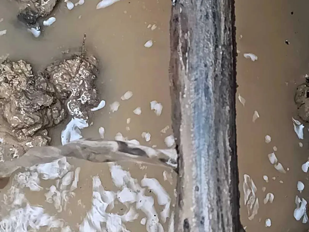Corroded metal supply pipe in a flooded hole, with a jet of water spraying from it into the muddy water