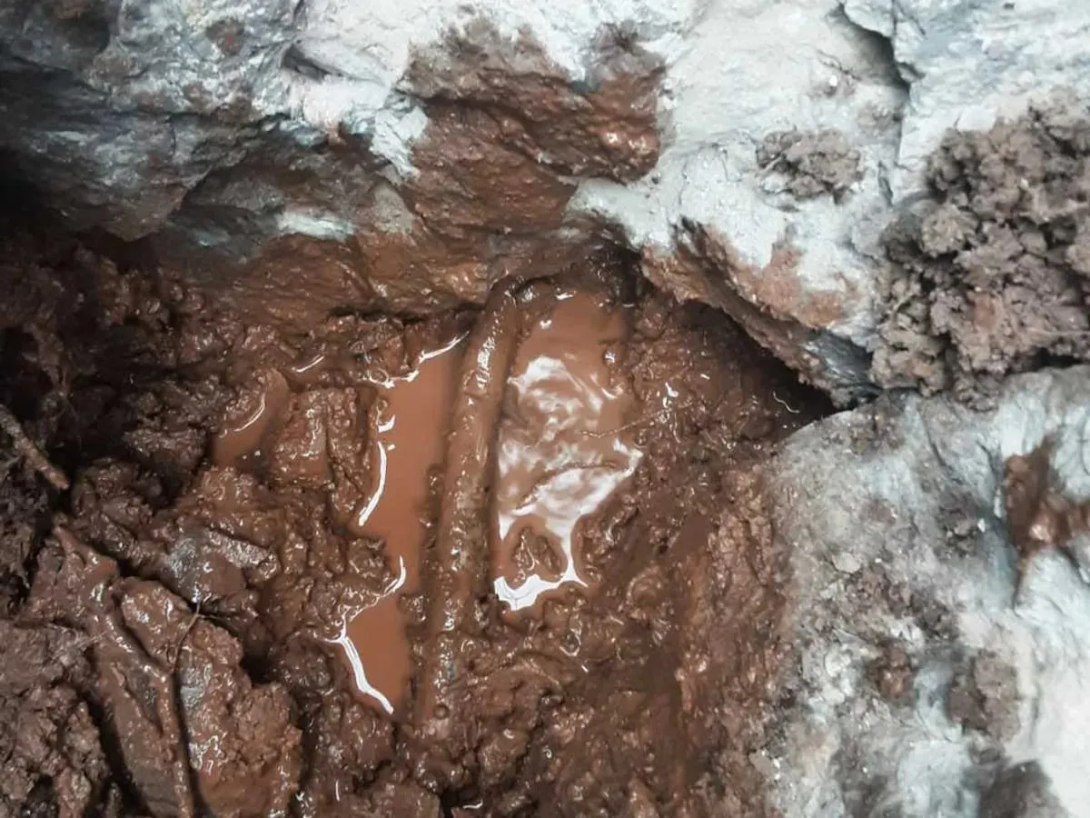 Muddy, waterlogged hole with the old supply pipe lying in brown water