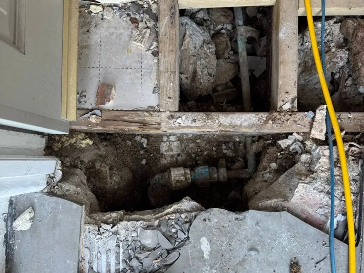 Threshold opened up with floor joists visible and the supply pipe and fittings exposed in the broken-out concrete below